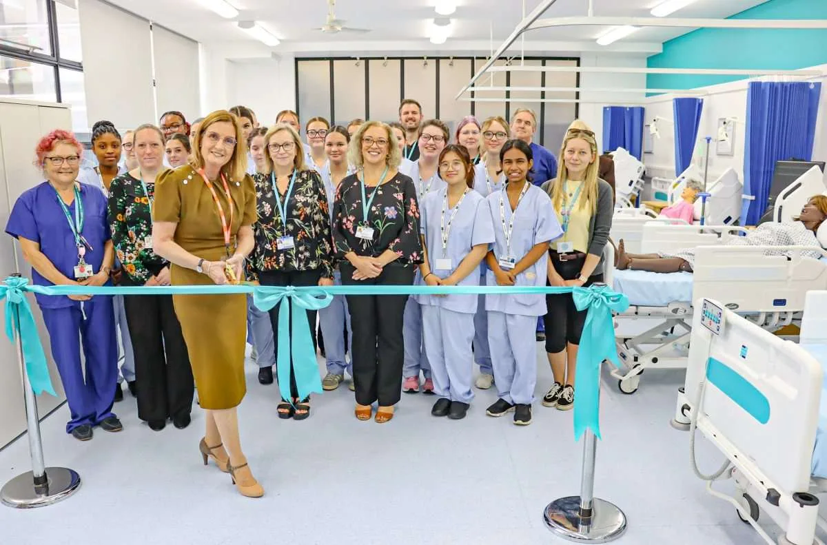 A group of college staff, hospital trust staff and students in pale blue scrubs stand behind a teal ribbon in a bright hospital-style room with beds and blue curtains, as the ribbon is cut.